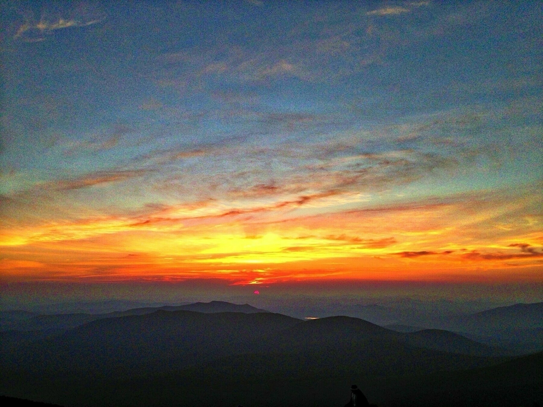 A vibrant sunrise over a mountainous landscape. The sky is filled with shades of orange, yellow, and hints of blue and purple, creating a dramatic contrast with the dark silhouettes of the rolling hills below. Soft, wispy clouds stretch across the sky, adding texture to the scene.