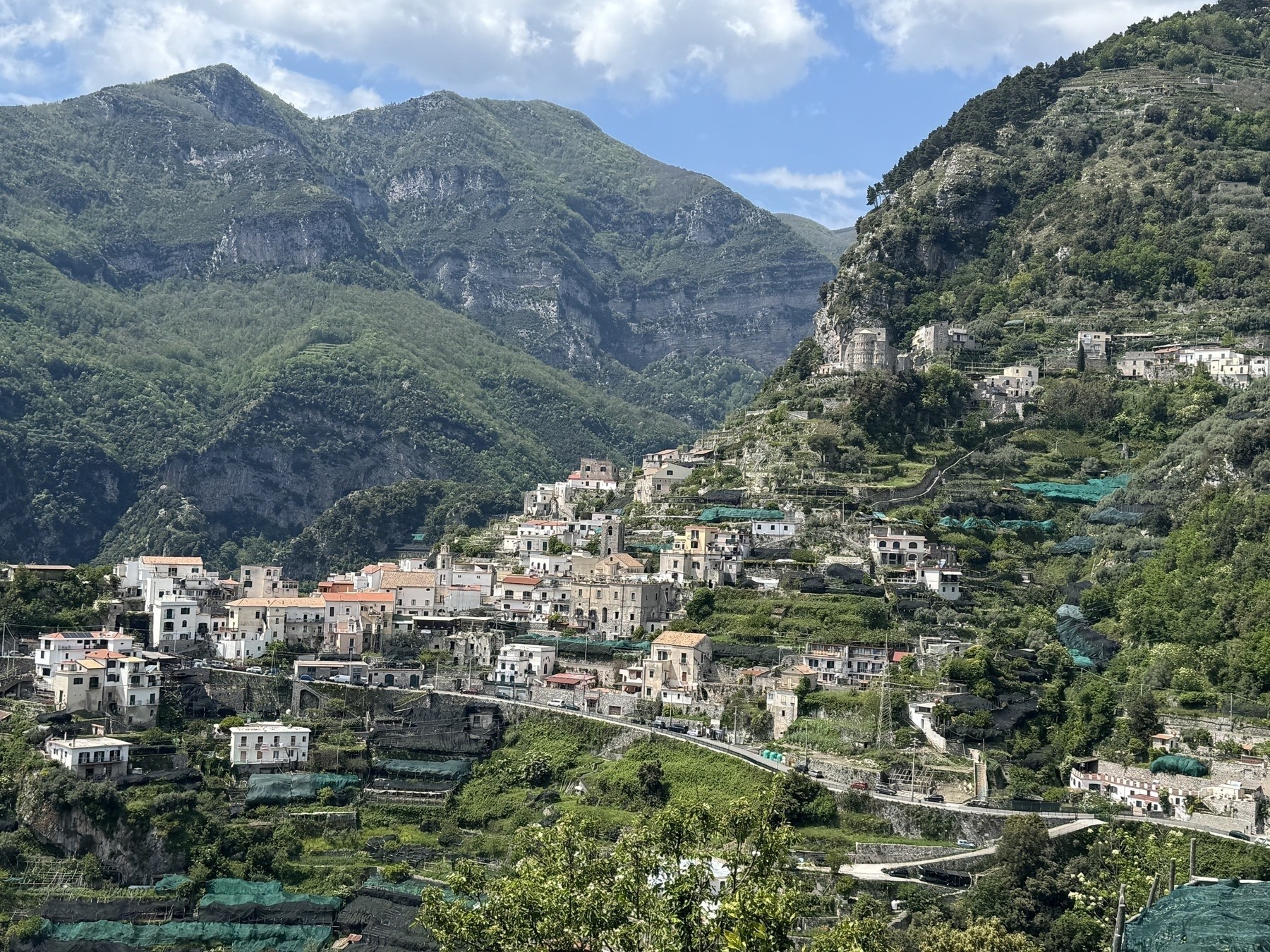 A picturesque hillside village nestled against a backdrop of lush green mountains. The scene features numerous buildings, mostly white with red-tiled roofs, scattered along terraced slopes. Verdant vegetation, including trees and bushes, surrounds the structures, with hints of cultivated fields interspersed throughout. The sky is partly cloudy with patches of blue visible. A picturesque hillside village nestled against a backdrop of lush green mountains. The scene features numerous buildings, mostly white with red-tiled roofs, scattered along terraced slopes. Verdant vegetation, including trees and bushes, surrounds the structures, with hints of cultivated fields interspersed throughout. The sky is partly cloudy with patches of blue visible.