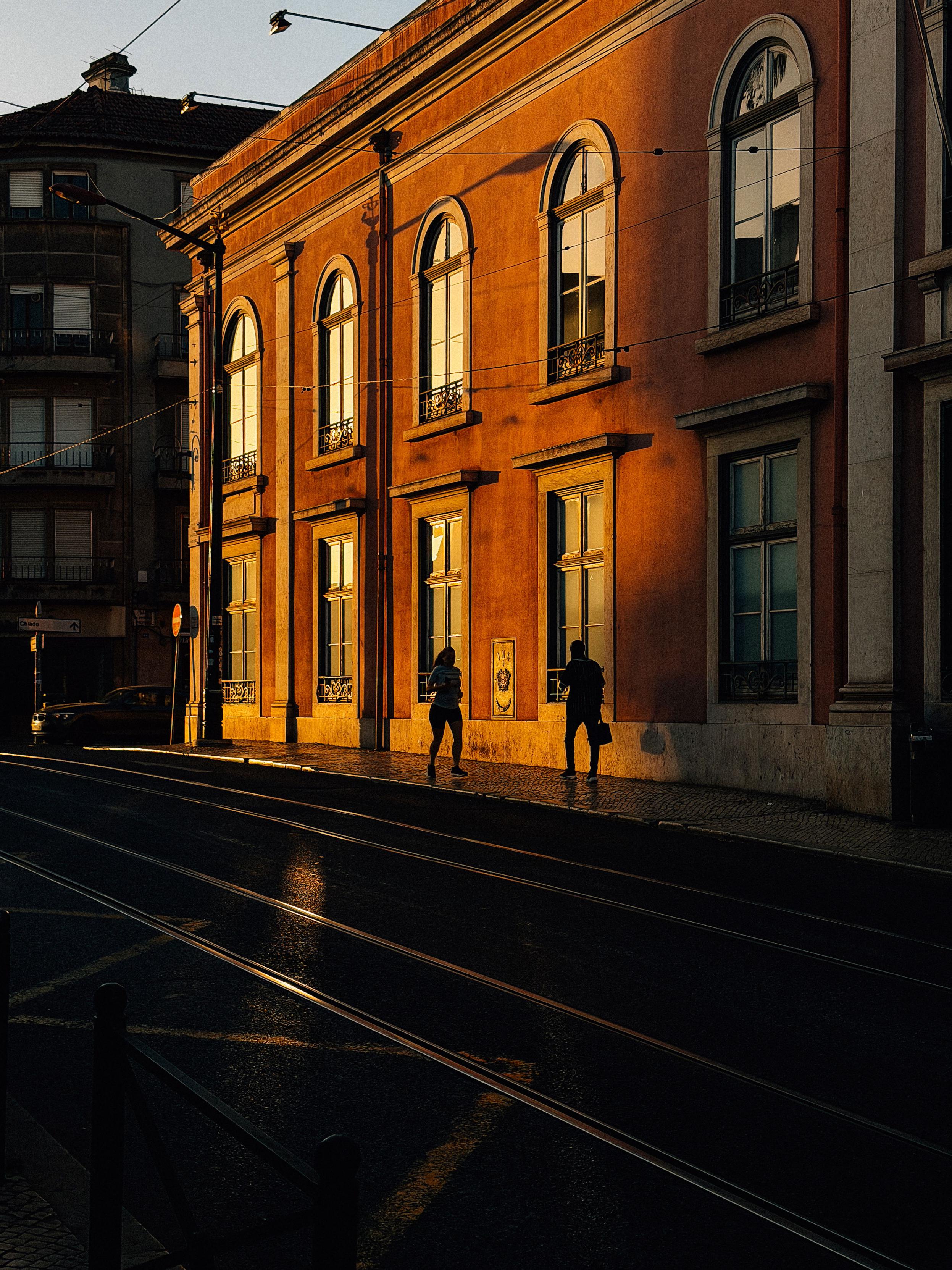Two people walking, against a classic looking building, in the morning light.
