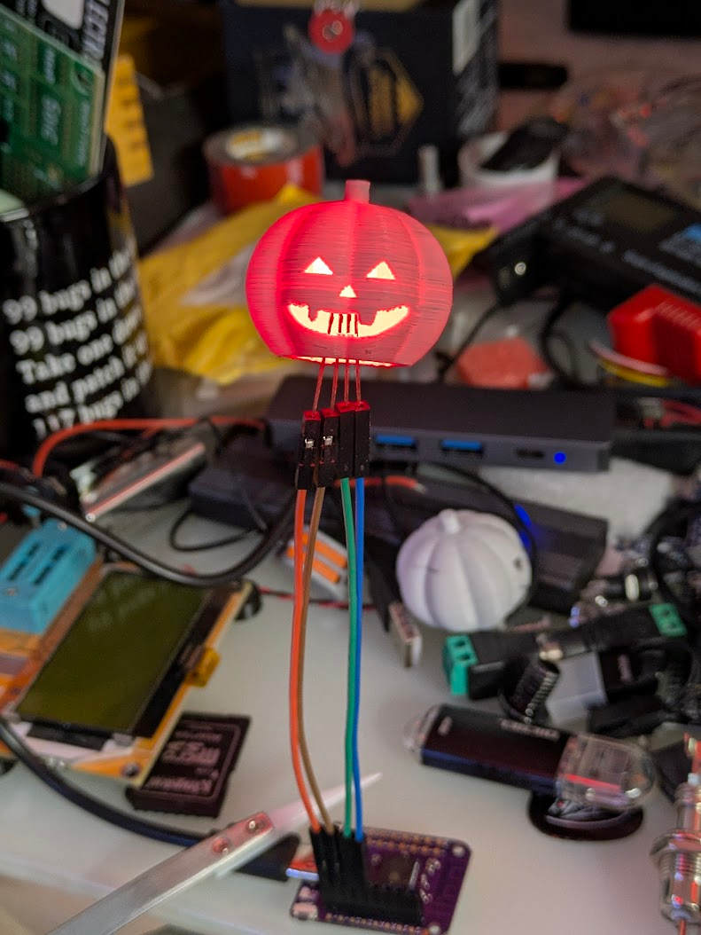a 3d printed jack-o-lantern that is glowing red from a single large RGB LED inside that is connected with dupont breadboard wires to an ESP32 S3 module, in the background there is all kinds of clutter on my messy desk