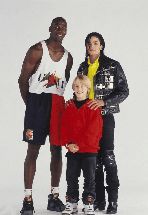 Three people stand together in a studio-style photo with a plain white background. On the left is a very tall, athletic-looking man (Michael Jordan) wearing a sleeveless sports jersey and basketball shorts. On the right is a man with long black hair, dressed in a shiny black jacket and black pants with decorative straps (Michael Jackson). Between them is a smiling blond child in a red sweatshirt (McCauley Culkin). The two adults rest their hands gently on the child’s shoulders.