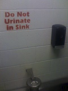 An old fashioned multi-person spray sink and above on the wall there is red stencil painted text:
Do Not
Urinate
in Sink